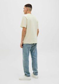 Lichtbeige T-shirt met een relaxte pasvorm, gecombineerd met lichtblauwe denim jeans. Model staat met de rug naar de camera en toont een casual stijl. Witte sneakers.