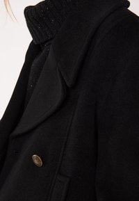 Cappotto di lana nero con ampio risvolto, dotato di un bottone decorativo dorato. La trama sembra morbida e spessa, adatta per il clima più freddo.