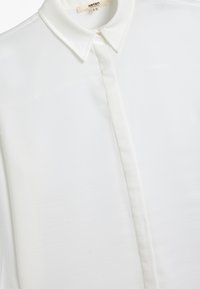 Camicia bianca con colletto, realizzata in tessuto liscio. Presenta una patta frontale e un design pulito e minimalista.