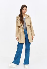 Beżowy lekki trench, z kapturem, z paskiem w talii, zamykanymi kieszeniami i wielowarstwowym przodem, w zestawie z paskowanym topem i niebieskimi dżinsami.