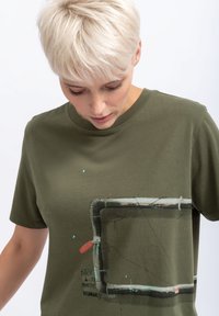 T-shirt vert olive en coton, avec un design graphique représentant un cadre de tuyau accompagné de texte et d'éclaboussures de couleur. Manches courtes, col rond.