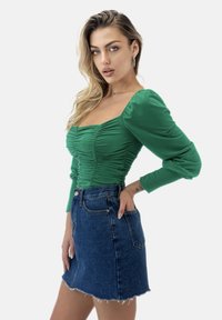 Top verde a maniche lunghe con dettagli arricciati e scollo quadrato abbinato a una mini gonna di jeans blu con orlo sfilacciato e tasche.