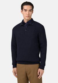 Tmavě modrý pletený svetr s límcem a třemi knoflíky, doplněný o tmavé tričko pod ním. Detail v žebrovaných manžetách a lemu.