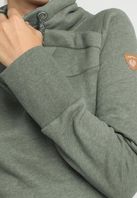Sudadera verde con cuello alto, tela texturizada y un bolsillo lateral. Presenta un cordón y un parche de la marca en la manga.