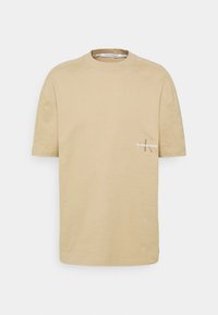 Camiseta de algodón beige con cuello redondo y mangas cortas, que presenta un pequeño logo bordado en el lado izquierdo del pecho. Textura suave.