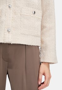 Veste texturée crème avec un tissage doux, fermeture à boutons en argent et une poche à rabat à l'avant, assortie à un pantalon marron.