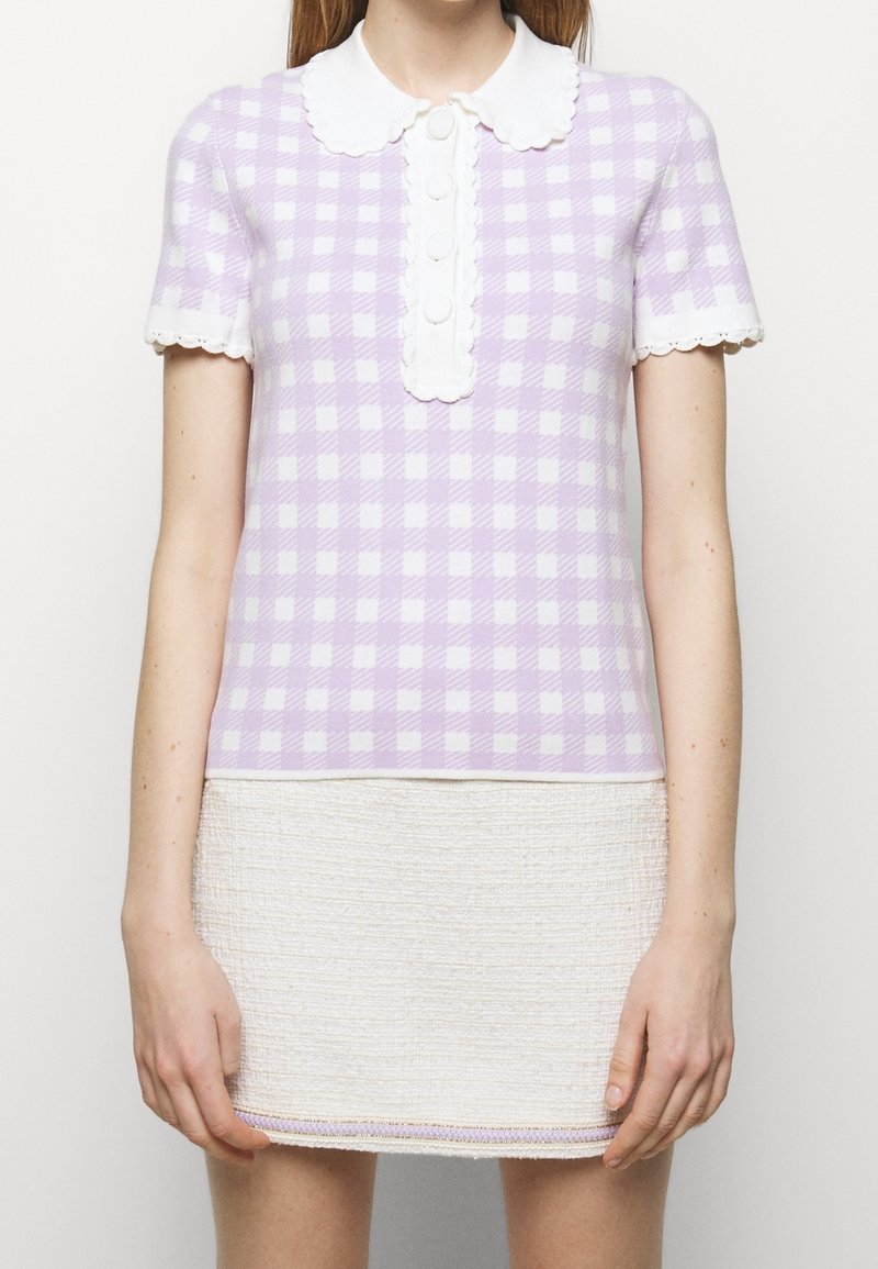 Kobieta w lawendowej i białej kratkowanej bluzce z krótkim rękawem, z białym, falbaniastym kołnierzykiem i guzikami, w zestawieniu z teksturowaną, kremową spódnicą.