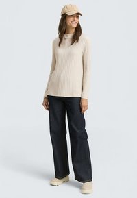 Šviesiai smėlio spalvos ribb megztinis ilgomis rankovėmis derinamas su tamsiais platėjusių kojų džinsais ir šviesiai smėlio spalvos platforminiu batais, papildytas rudos spalvos beisbolo kepuraitė.