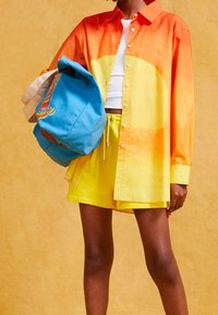 Oversized overhemd in een verloop van oranje en geel, gecombineerd met gele korte broek. Houdt een blauwe rugzak vast met kleurrijke borduursels.