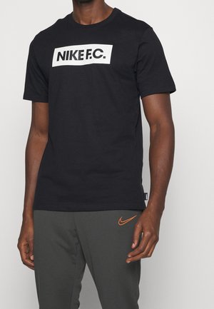 Mężczyzna w czarnym t-shircie Nike F.C. i ciemnoszarych spodniach z pomarańczowym logo Nike swoosh na lewym udzie.