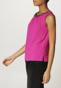 Rosa ärmlös topp med rundad halsringning som har en dekorativ, pärlbesatt krage. Tyget ser slätt ut; sidoklipp vid fållen.