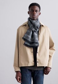 Jachetă impermeabilă bej, eșarfă neagră cu modele de logo, blugi slim; textură netedă, croială casual, butoane frontale și detalii cu franjuri pe eșarfă.