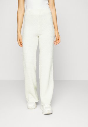 Pantalons blancs, texturés, à jambes larges avec une taille épaisse et côtelée, associés à des baskets blanches. Le tissu semble doux et duveteux.