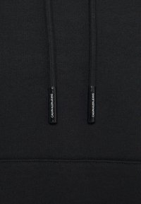 Sudadera con capucha negra de tejido texturizado, que presenta cordones negros con herrajes plateados y la marca "Calvin Klein Jeans".