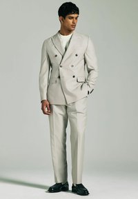 Suit grigio chiaro doppiopetto con bottoni neri, design slim-fit e pantaloni con frontale piatto. Abbinato a una camicia bianca a girocollo e scarpe nere.