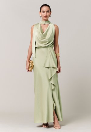 Ženska, oblečena v brezrokavno svetlo zeleno satenasto obleko z nagubanim stranskim detajlom, zlato zapestnico, zlato torbico in zlatimi sandali na enobarvnem ozadju.