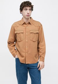 Καφέ πουκάμισο με μακριά μανίκια, δύο τσέπες στο στήθος, κουμπιά μπροστά και αντίθετες ραφές. Διακριτική λεπτομέρεια με θηλιά κοντά στο τελείωμα, συνοδευόμενο από μπλε τζιν.