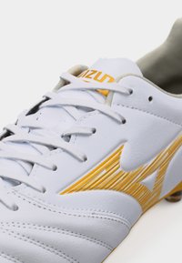 Λευκό δερμάτινο παπούτσι ποδοσφαίρου με κίτρινο λογότυπο Mizuno, λευκά κορδόνια και ραφές στην περιοχή των δακτύλων.