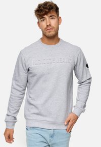Sudadera de color gris claro con puños y dobladillo acanalados, con "INDICODE JEANS" grabado en la parte delantera y un pequeño logo negro en la manga.