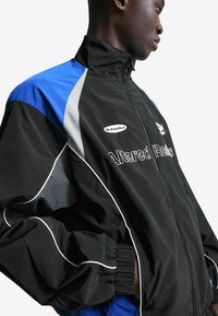 Man in een zwart, blauw en grijze trainingsjack met witte piping, staande met zijn handen in zijn zakken tegen een witte achtergrond.