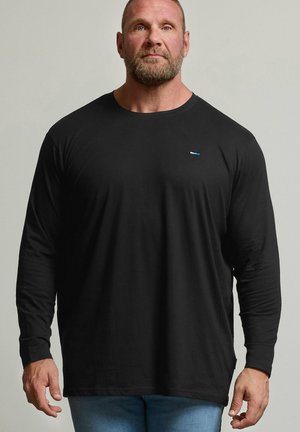 Mann mit Bart, der ein schlichtes schwarzes Langarmshirt und hellblaue Jeans trägt, steht nach vorne gerichtet vor einem neutralen Hintergrund.