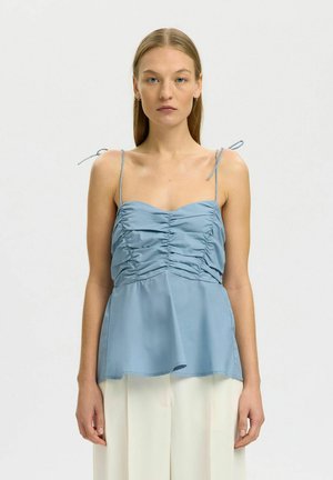 Femme portant un haut sans manches bleu clair froncé avec de fines bretelles aux épaules et un pantalon blanc à jambes larges, debout devant un fond uni.