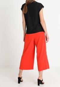 Svart kortärmad topp med texturerat tyg, kombinerad med ljusröda vida byxor. Modellen har på sig svarta klacksandaler.