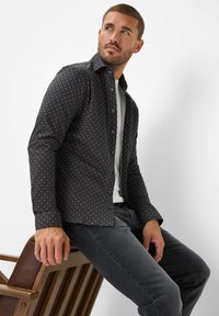 Camicia a maniche lunghe in tessuto scuro con un motivo geometrico, colletto a punta e chiusura a bottoni, abbinata a pantaloni grigi.