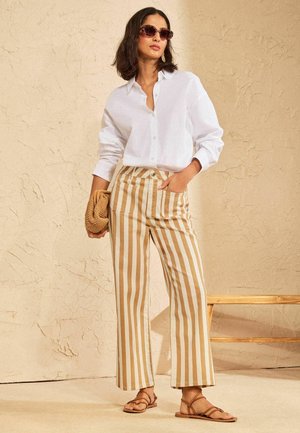 Vrouw draagt een witte blouse met knopen, beige en wit gestreepte wijde broek, bruine sandalen, zonnebril en houdt een gevlochten clutch vast.
