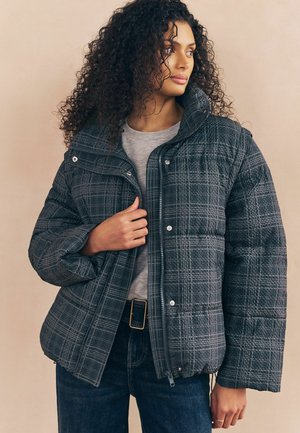 Vrouw met krullend haar die een grijze geruite pufferjack draagt over een grijs T-shirt en donkere jeans, poserend voor een beige achtergrond.