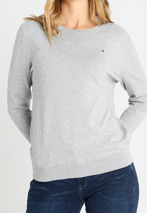 Šedý pletený svetr s kulatým výstřihem, dlouhými rukávy a malým logem na hrudi. Jemná textura, kombinován s tmavě modrými džínami.