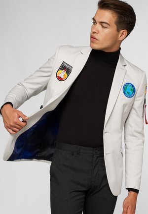 Blazer bianco con toppe a tema spaziale; include un dolcevita nero e pantaloni grigi. Foderato con un tessuto stampato galassia.