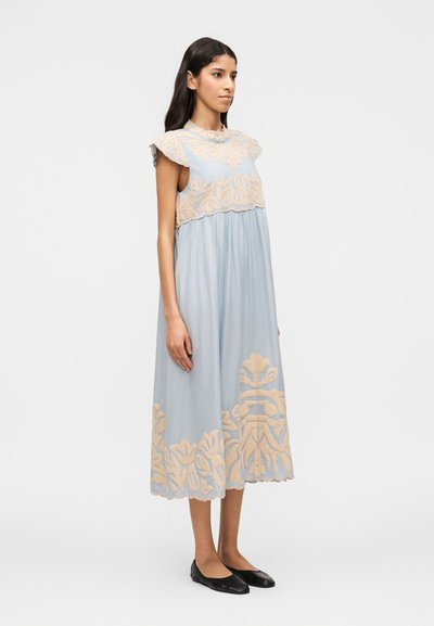 Γυναίκα που φοράει ένα χαλαρό, αμάνικο φωτεινό μπλε φόρε μα με μπεζ λουλουδάτη κεντητική, που στέκεται μπροστά σε απλό λευκό φόντο.