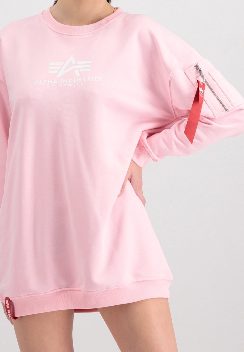 Różowa bluza o luźnym kroju, z logo na piersi, kieszenią na zamek na rękawie oraz czerwonym akcentem na metce przy dolnym brzegu.