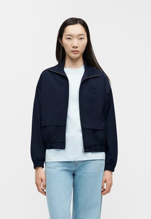Giovane donna con lunghi capelli scuri che indossa una giacca blu navy con zip sopra una camicia azzurra e jeans azzurri, in piedi su uno sfondo semplice.