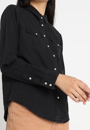 Femme portant une chemise noire boutonnée avec des pressions en perle et un pantalon beige, présentée de la poitrine aux hanches sur fond blanc.
