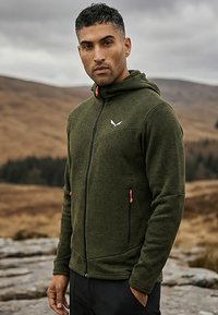 Zöld, cipzáros kapucnis dzsekit viselő férfi, narancssárga cipzárhúzókkal, szabadtéren, sziklás terepen áll, háttérben dombok és felhős égbolt.