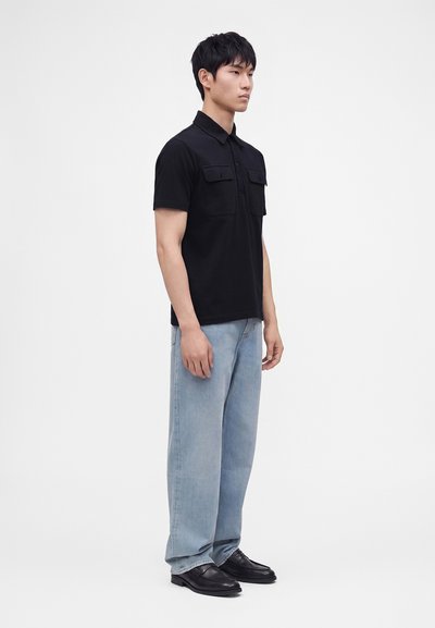 Homme debout portant une chemise noire à manches courtes avec poches poitrine, un jean bleu clair et des chaussures en cuir noires sur fond blanc uni.