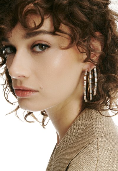 Mlada ženska s kodrastimi lasmi, ki nosi velike trojne obročaste uhane in teksturirano bež blazer jakno, rahlo obrnjena vstran.