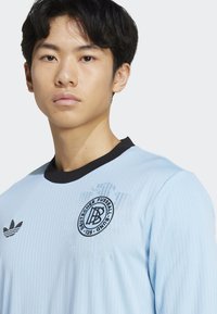Lichtblauw overhemd met lange mouwen en een zwart geribbeld kraagje, met een rond logo op de linkerkant van de borst en subtiele verticale strepen verspreid over het hele shirt.