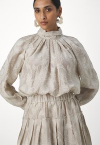 Bluză bej brodată cu gât înalt și mâneci bufante; material texturat cu modele florale; talie strânsă pentru o siluetă fluidă.