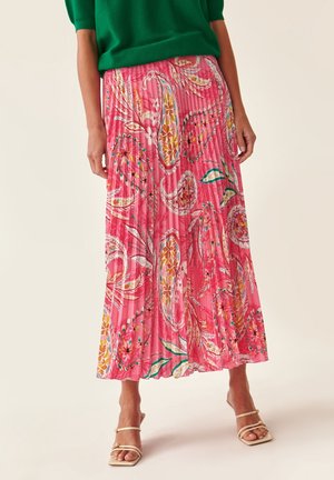 Woman wearing a green top and a pleated pink floral maxi skirt with white strappy heels, standing against a plain background.