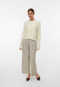 Cardigan a maglia di colore crema con texture orizzontale, chiuso da bottoni dorati. Abbinato a pantaloni larghi beige e scarpe marroni a punta.