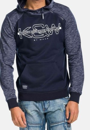 Homme portant un sweat à capuche bleu marine avec des manches grises et un jean bleu clair, texte sur le devant du sweat à capuche : « KSW Koroshi Worldwide Denim Lab by MNKB. »