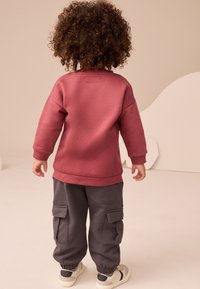 Vaikas dėvi bordo spalvos džemperį su gofruotomis rankogaliais ir apatinėmis kraštinėmis, derinamas su tamsiai pilkomis kargo kelnėmis su šoniniais kišenėmis.