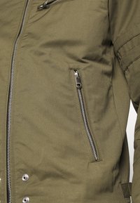 Nära bild av en olivgrön jacka med lodrätt silverfärgad dragkedjeficka och huvuddelen av dragkedjan delvis synlig.