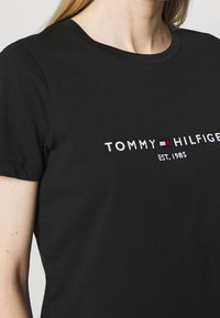 Žena nosící černé tričko s vyšívaným nápisem "Tommy Hilfiger" v bílé barvě a vlajkovým logem značky v červené, bílé a modré barvě na prsou.