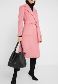 Kvinde iført rosa bæltejakke, sort turtleneck, sorte bukser og sok-støvler, der holder en stor sort læder Coach-handtaske.