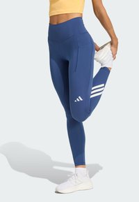 Leggings bleu taille haute avec une texture lisse, dotés d'une poche latérale et d'un design à trois bandes blanches sur la partie inférieure de la jambe, assortis à des chaussures de sport blanches.