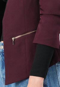 Veste bordeaux avec une poche zippée, texture de tissu lisse, manches larges et un sous-pull noir côtelé visible au poignet.